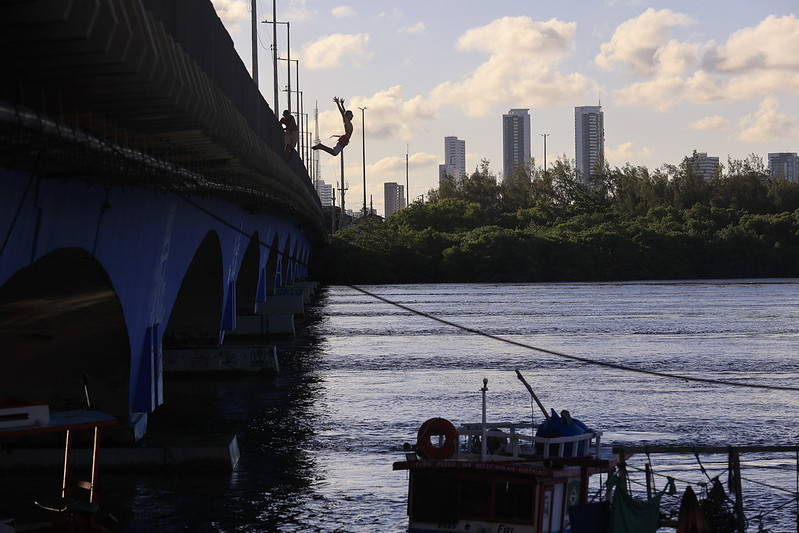 Na imagem, vemos um jovem saltando de uma ponte em direção ao rio, na Bacia do Pina, no Recife. O corpo dele está inclinado para trás, com os braços erguidos e as pernas dobradas, no momento exato em que está no ar. A ponte, pintada de azul em sua parte inferior, ocupa o lado esquerdo da foto. À frente, a água reflete a luz do céu, que está parcialmente nublado com nuvens claras. No fundo, aparecem prédios altos da cidade, atrás de uma faixa de árvores verdes. Na parte inferior direita da imagem, há um pequeno barco de pesca com boias e redes penduradas.