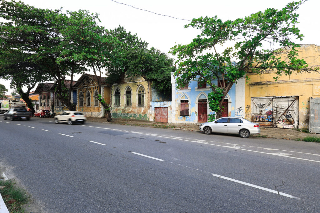 A foto mostra uma rua urbana com prédios de vários andares em arquitetura antiga, com sacadas e fachadas em tons claros e detalhes ornamentais sutis. No centro da via, há uma van escura estacionada, e ao fundo a rua se eleva em uma ladeira que dá profundidade à cena. As calçadas aparecem desgastadas, com sinais de uso ou reparos. A iluminação é de um dia claro, com luz natural incidindo sobre os edifícios. Também se observam fios elétricos expostos e postes que reforçam o ambiente típico de uma área urbana.