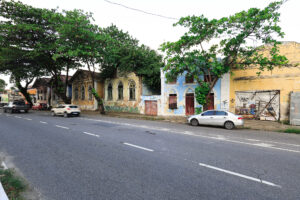 A foto mostra uma rua urbana com prédios de vários andares em arquitetura antiga, com sacadas e fachadas em tons claros e detalhes ornamentais sutis. No centro da via, há uma van escura estacionada, e ao fundo a rua se eleva em uma ladeira que dá profundidade à cena. As calçadas aparecem desgastadas, com sinais de uso ou reparos. A iluminação é de um dia claro, com luz natural incidindo sobre os edifícios. Também se observam fios elétricos expostos e postes que reforçam o ambiente típico de uma área urbana.