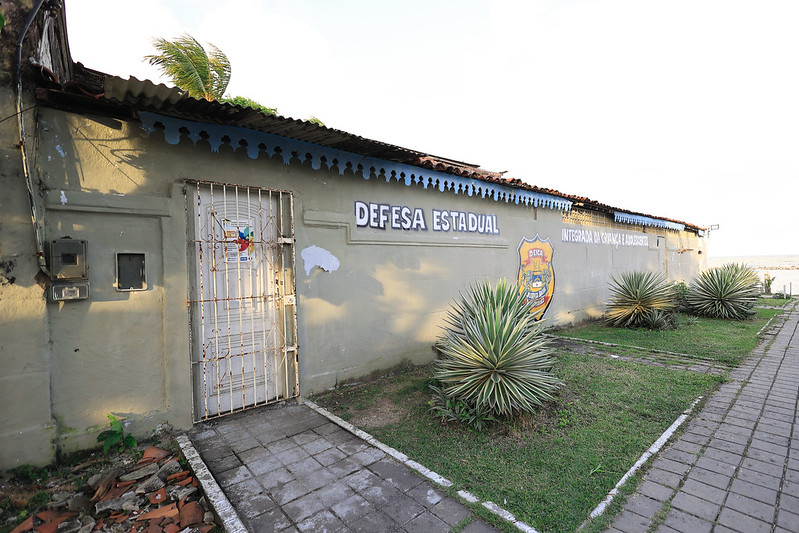 A foto mostra a fachada simples de um prédio baixo, pintado em tom claro. Na parede, em destaque, lê-se em letras maiúsculas “DEFESA ESTADUAL”, acompanhado de um brasão institucional. A porta de entrada é metálica, branca e gradeada, com fechadura visível. Em frente, há um pequeno jardim com plantas ornamentais de folhas pontiagudas sobre solo gramado. À direita, um caminho calçado leva até a porta. O telhado tem beiral de telhas cerâmicas, mostrando sinais de desgaste. Uma árvore aparece à esquerda, projetando sombra sobre parte da construção. O céu ao fundo está claro, com algumas nuvens leves. A cena transmite simplicidade e solidez de um espaço oficial.