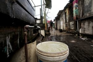 A imagem mostra uma cena em uma comunidade popular, possivelmente em uma área de palafitas. Em primeiro plano, há um balde branco sendo enchido com água de uma torneira simples fixada a uma parede de madeira escura. A água que cai parece turva, sem transparência. O chão é de tábuas de madeira molhadas, dando a impressão de precariedade e risco. Ao fundo, é possível ver casas feitas de madeira e metal, com algumas paredes pintadas com grafites coloridos. Algumas pessoas aparecem caminhando pelo beco estreito, uma delas segurando um guarda-chuva vermelho.