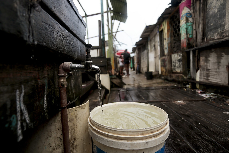 A imagem mostra uma cena em uma comunidade popular, possivelmente em uma área de palafitas. Em primeiro plano, há um balde branco sendo enchido com água de uma torneira simples fixada a uma parede de madeira escura. A água que cai parece turva, sem transparência. O chão é de tábuas de madeira molhadas, dando a impressão de precariedade e risco. Ao fundo, é possível ver casas feitas de madeira e metal, com algumas paredes pintadas com grafites coloridos. Algumas pessoas aparecem caminhando pelo beco estreito, uma delas segurando um guarda-chuva vermelho.