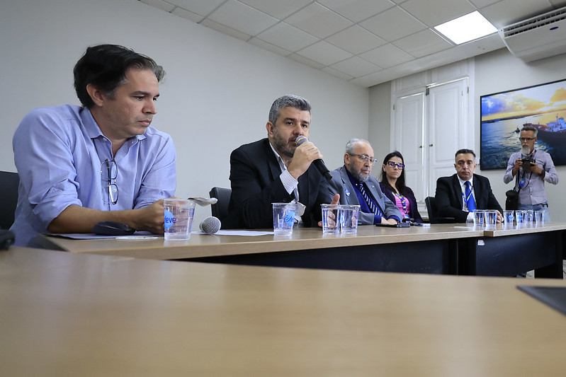 A imagem mostra uma entrevista coletiva realizada em uma sala de reuniões. Seis pessoas estão sentadas lado a lado atrás de uma mesa comprida de madeira clara. No centro, um homem de terno preto fala ao microfone, aparentando ser o porta-voz ou principal representante do grupo. À sua esquerda, outro homem de óculos, também de terno, observa atentamente, enquanto ao lado dele uma mulher de óculos, de roupa roxa, acompanha a fala. À direita do porta-voz, há dois homens em trajes formais, igualmente atentos. À extrema esquerda da mesa, um homem de camisa social azul clara está com os braços apoiados, olhando para baixo. Sobre a mesa, há copos plásticos com água e dois microfones. Ao fundo, a parede é clara com uma porta de madeira e um quadro com uma fotografia colorida de um pôr do sol sobre o mar. À direita, um homem em pé segura uma câmera, registrando a coletiva.