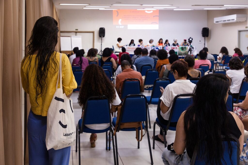 A imagem mostra uma sala com várias pessoas sentadas em cadeiras azuis, assistindo a um painel de discussão. Na frente, há uma mesa longa com palestrantes, coberta por bandeiras. Um telão exibe uma apresentação, embora o texto não esteja legível. Uma pessoa em pé, ao fundo, veste blusa amarela e calça azul, carregando uma bolsa branca com a palavra “labor”.