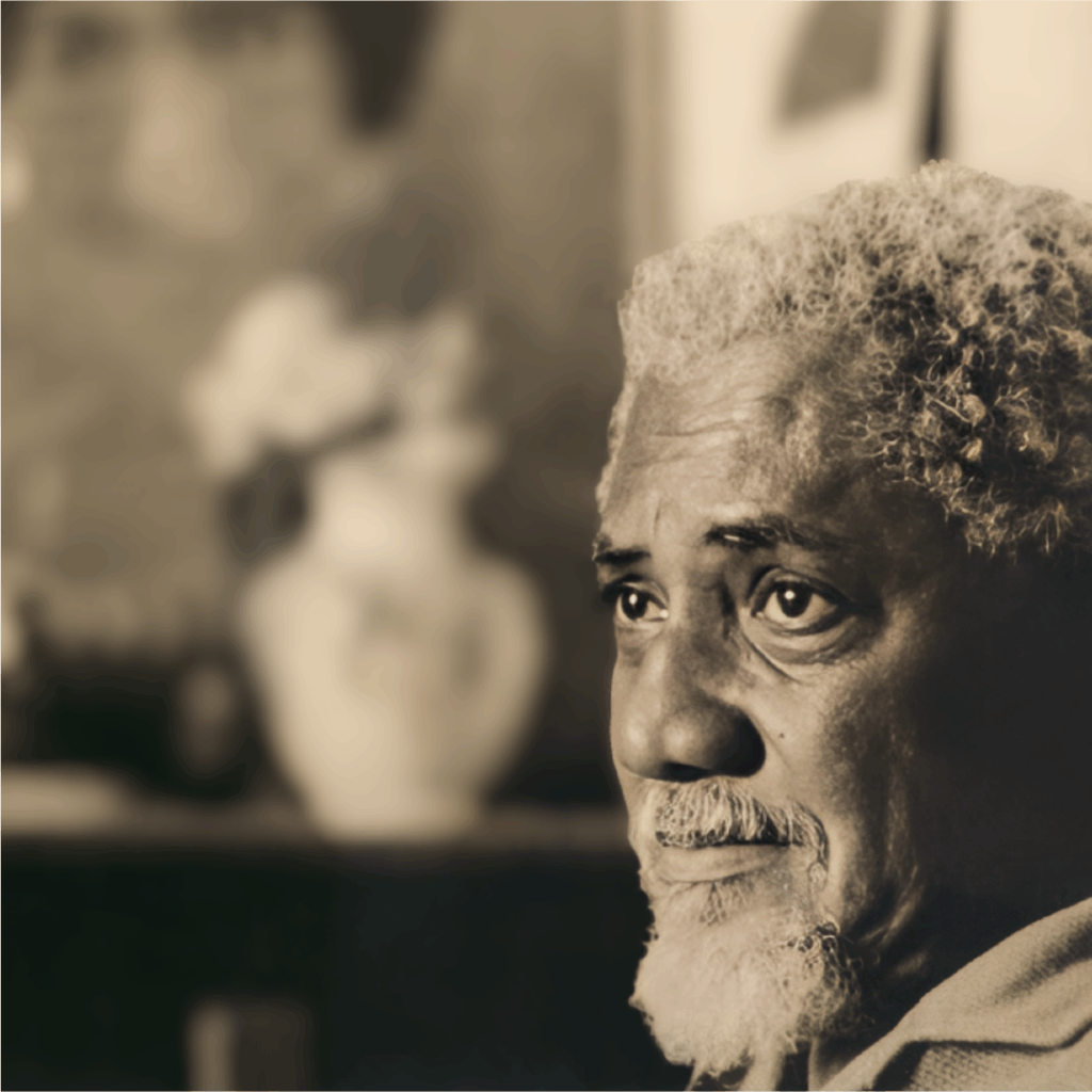 A imagem mostra o rosto de um homem negro idoso, com cabelo grisalho e encaracolado e barba. Ela olha pensativa para o lado, com expressão introspectiva. Ao fundo, há um vaso branco com flores e quadros desfocados na parede. A luz destaca os traços do rosto, criando um clima íntimo e sereno. É um retrato que transmite contemplação em um ambiente acolhedor.