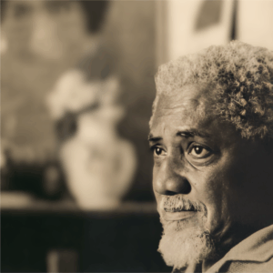 A imagem mostra o rosto de um homem negro idoso, com cabelo grisalho e encaracolado e barba. Ela olha pensativa para o lado, com expressão introspectiva. Ao fundo, há um vaso branco com flores e quadros desfocados na parede. A luz destaca os traços do rosto, criando um clima íntimo e sereno. É um retrato que transmite contemplação em um ambiente acolhedor.