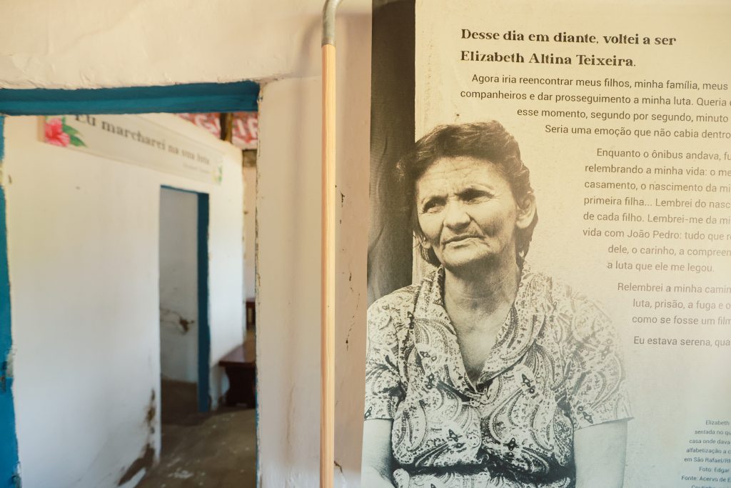Imagem em preto e branco de Elizabeth Altina Teixeira, uma mulher com rosto marcado e expressão séria ou pensativa, olhando ligeiramente para o lado direito da foto. Ela veste uma blusa estampada com um padrão floral ou de folhagens. A foto é parte de um painel de exibição. À direita dela, há um texto em português que começa com a frase: Desse dia em diante, voltei a ser Elizabeth Altina Teixeira. À esquerda da imagem, um vão de porta ou parede interna, pintada de branco com a borda emoldurada em azul e um pedaço de folha de papel com a frase Eu marcharei na tua luta visível. Um pedaço de madeira vertical e fino separa o painel do vão de porta.