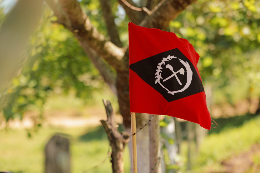 Close de uma bandeira vermelha em tecido, presa a uma pequena estaca de madeira e ligeiramente ondulada. No centro da bandeira, há um losango (forma de diamante) preto, e dentro dele, um círculo branco com um desenho de um machado cruzado com uma foice. A bandeira está em um ambiente externo, à luz do sol, com o fundo desfocado mostrando árvores, galhos e folhagens verdes.