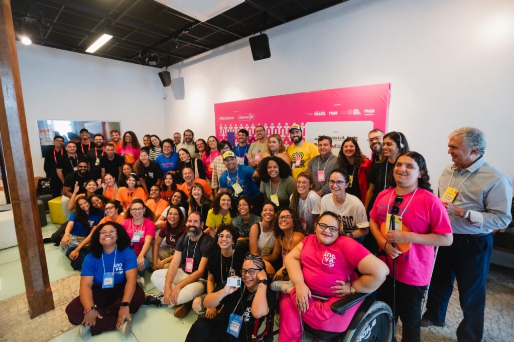 Foto colorida de um grande grupo de pessoas reunidas em um espaço interno, em frente a um painel rosa com a palavra “inova aê”. A maioria sorri para a câmera, algumas sentadas no chão, outras em pé. O grupo é diverso em idades, estilos e inclui pessoas com deficiência. O clima é de alegria e celebração coletiva.