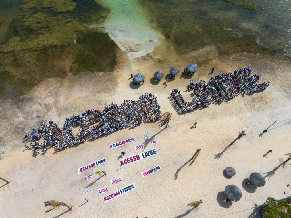 A imagem mostra uma manifestação no Parque Nacional de Jericoacoara, no Ceará, vista de cima. Centenas de pessoas estão reunidas na areia clara, próximas ao mar, formando com os próprios corpos a frase “ACESSO LIVRE”. Logo abaixo, há várias faixas estendidas no chão com mensagens como “#ACESSO LIVRE”, “ACESSO LIVRE”, “#JERISempreLivre”, “#JERI NÃO É PARQUE” e “NÃO À CONCESSÃO”. O cenário natural inclui o mar esverdeado à esquerda, algumas palmeiras espalhadas e pequenos quiosques de palha que fazem sombra. A manifestação parece organizada para defender o livre acesso à praia e protestar contra restrições ou concessões dentro do parque.