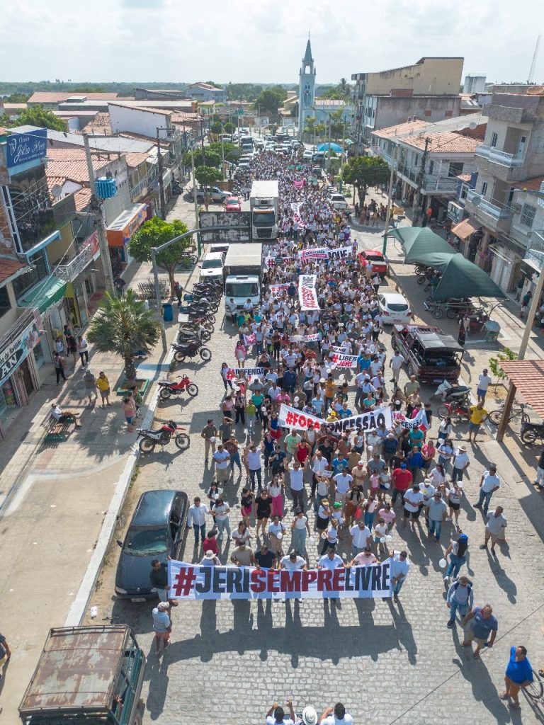A imagem mostra uma manifestação de rua em Jericoacoara. Pessoas de diferentes idades estão reunidas ao longo de uma via ou calçada, segurando cartazes, faixas e bandeiras. Muitas carregam plaquinhas com mensagens, provavelmente cobrando “acesso livre”, com reivindicações relacionadas ao uso do parque nacional ou à cobrança de taxas. Há um sentimento de mobilização comunitária, expressado no corpo a corpo — manifestantes amontoados, alguns com as mãos erguidas, interagindo uns com os outros.