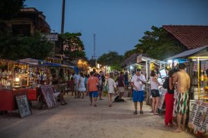 A foto mostra uma rua movimentada da vila de Jericoacoara, no Ceará, durante o início da noite. O chão é de areia clara e há várias barracas iluminadas dos dois lados da rua, vendendo bebidas, artesanatos e comidas típicas. Algumas placas e cardápios escritos à mão estão expostos em frente às barracas. Pessoas caminham de forma descontraída, muitas com roupas leves de verão, shorts, camisetas e saídas de praia, algumas descalças. Há também pequenos grupos conversando e pessoas fazendo pedidos nos quiosques. O clima é de descontração e turismo, com iluminação aconchegante das luzes das barracas contrastando com o céu azul-escuro do entardecer. Ao fundo, é possível ver casas e comércios com fachadas rústicas, árvores e vegetação completando a cena típica de uma vila litorânea.
