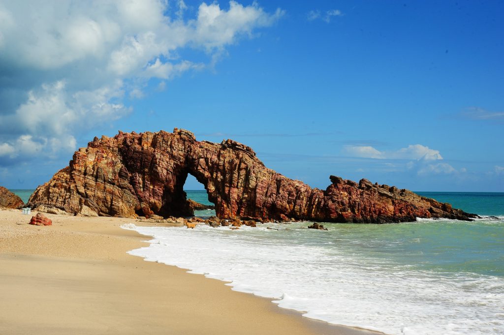 A foto mostra a Pedra Furada, um dos cartões-postais mais famosos de Jericoacoara, no Ceará. Trata-se de uma formação rochosa natural à beira-mar, com um grande arco no centro, moldado pela ação do tempo e do mar. As rochas têm tons avermelhados, amarelados e marrons, criando um contraste marcante com o azul do céu e do oceano. O mar avança suavemente sobre a areia clara da praia, formando uma fina faixa de espuma branca. Ao fundo, o horizonte se mistura entre o azul do mar e as nuvens brancas no céu parcialmente aberto.
