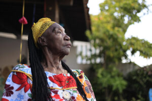 A imagem mostra a artista Lia de Itamaracá, uma mulher idosa de pele negra, com longos cabelos trançados. Ela veste um vestido colorido com estampas grandes de flores vermelhas, laranjas, azuis e verdes, e usa no peito um colar com medalhão dourado. Na cabeça, traz um turbante ou lenço amarelo rendado. Lia aparece em meio perfil, olhando para o lado esquerdo da foto, com expressão serena e firme. Ao fundo, vê-se parte de uma casa simples com telhado de madeira e algumas plantas verdes iluminadas pela luz natural.