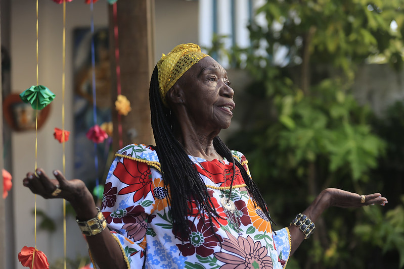 A imagem mostra a artista Lia de Itamaracá, uma mulher idosa de pele negra e cabelos longos trançados. Ela veste um vestido colorido com grandes estampas de flores em tons de vermelho, laranja, azul, verde e branco. Usa um turbante amarelo rendado na cabeça, pulseiras douradas nos braços e um colar com medalhão. Lia aparece sorridente, de braços abertos e levemente erguidos, em um gesto acolhedor. Ao fundo, há plantas verdes e algumas decorações coloridas de papel penduradas, que dão um ar festivo à cena.