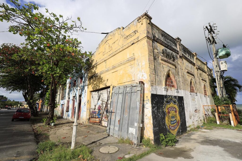 A fotografia mostra um prédio antigo, de esquina, com pintura amarelada desgastada e placas de madeira apoiadas contra a fachada. Algumas partes estão pichadas, inclusive um símbolo escuro com letras que parecem formar “DEICA”. O edifício tem janelas com formatos arqueados, parcialmente danificadas. Ao fundo, à esquerda, observa-se uma rua com calçada, árvores frondosas e um carro estacionado. No canto direito, há um poste de energia com transformador, e ao fundo direito, vislumbra-se o mar sob o céu parcialmente nublado.