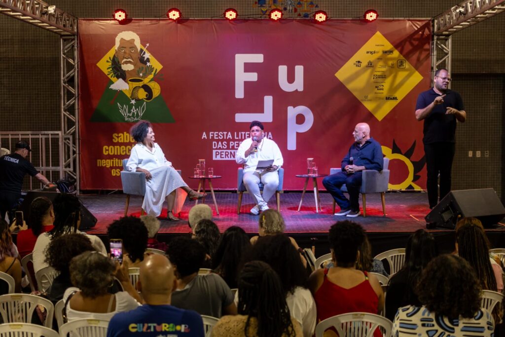 A foto mostra um evento cultural em um palco. Três pessoas estão sentadas em cadeiras de auditório no centro, participando de uma conversa: à esquerda, uma mulher de vestido claro; no meio, um homem de camisa branca; e à direita, um homem de roupa escura. Eles estão diante de um fundo vermelho com logotipos e o nome do evento escrito em destaque: “Flup – A Festa Literária das Periferias”. À direita do palco, um intérprete de Libras em pé traduz a fala dos participantes. Em frente ao palco, várias pessoas do público assistem sentadas em cadeiras de plástico brancas.