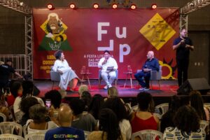 A foto mostra um evento cultural em um palco. Três pessoas estão sentadas em cadeiras de auditório no centro, participando de uma conversa: à esquerda, uma mulher de vestido claro; no meio, um homem de camisa branca; e à direita, um homem de roupa escura. Eles estão diante de um fundo vermelho com logotipos e o nome do evento escrito em destaque: “Flup – A Festa Literária das Periferias”. À direita do palco, um intérprete de Libras em pé traduz a fala dos participantes. Em frente ao palco, várias pessoas do público assistem sentadas em cadeiras de plástico brancas.