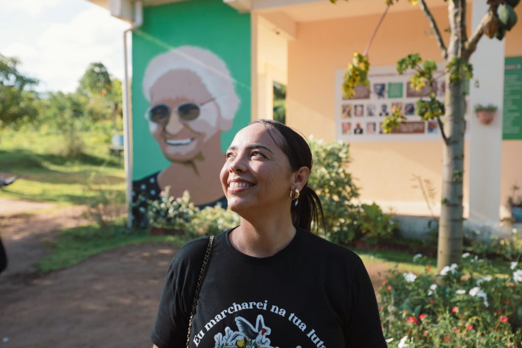A foto mostra Juliana Elizabeth Teixeira, uma mulher jovem de cabelos escuros, lisos e compridos, caindo pelos ombros. Ela aparece de perfil, levemente virada para o lado direito da imagem, com o olhar voltado na direção contrária à câmera. Sua expressão é serena, quase contemplativa. Veste uma camisa clara de mangas longas. O fundo é escuro ou desfocado, o que realça seu rosto e seus traços. A iluminação incide principalmente sobre o rosto, criando contraste com o cabelo e o entorno em sombra.