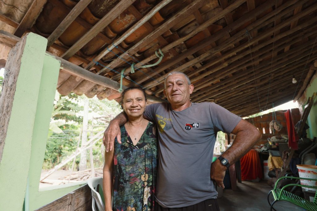 Um casal posa sorrindo para a câmera em um ambiente de varanda rústica. O homem, de pele clara e cabelo grisalho, veste uma camiseta cinza e abraça a mulher com o braço direito. A mulher, de pele morena, veste um vestido estampado com decote em V. Eles estão sob um telhado de madeira e telhas aparente. Há um pilar verde de alvenaria à esquerda. O fundo, atrás do casal, tem folhagens verdes e objetos de casa.