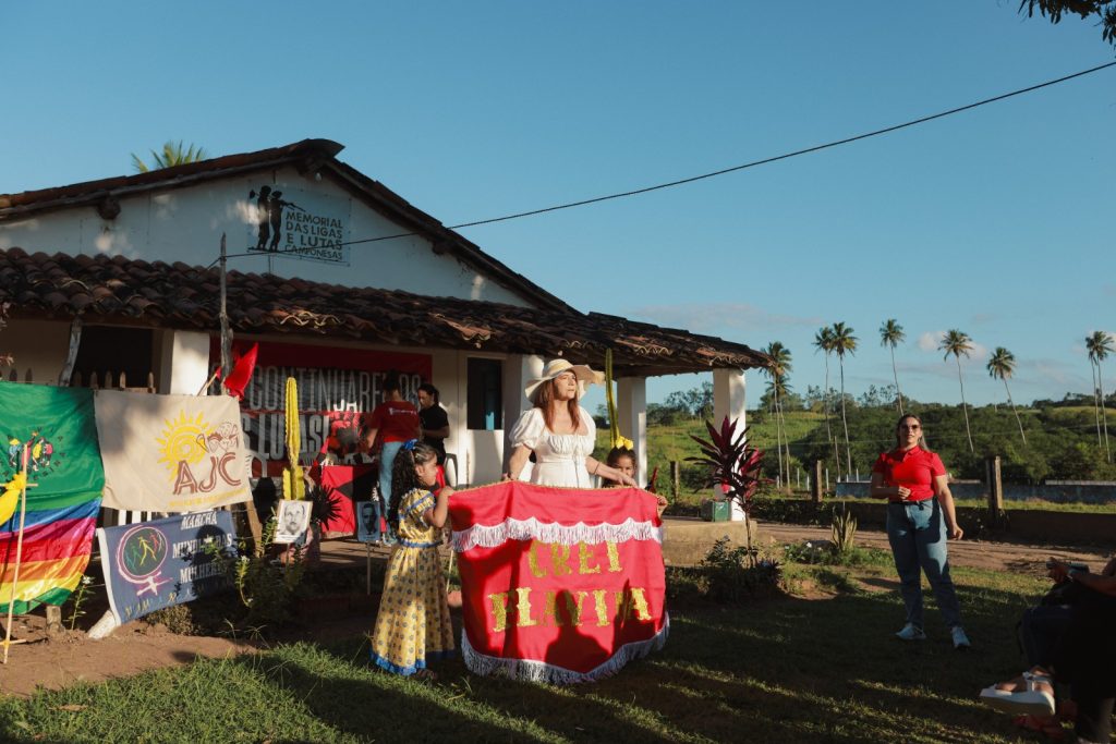 Cena externa de um evento ao ar livre, no final da tarde. Uma mulher com um vestido branco e um chapéu de palha está de pé no centro, à frente de uma construção baixa, simples e branca. Ela segura um pedaço de tecido grande, vermelho e amarelo, que tem uma inscrição, provavelmente um estandarte. No gramado ao lado, há outras pessoas e várias bandeiras coloridas e faixas, algumas com logotipos de organizações (como AJC). O fundo é um belo céu azul com coqueiros e um pequeno monte gramado no horizonte.