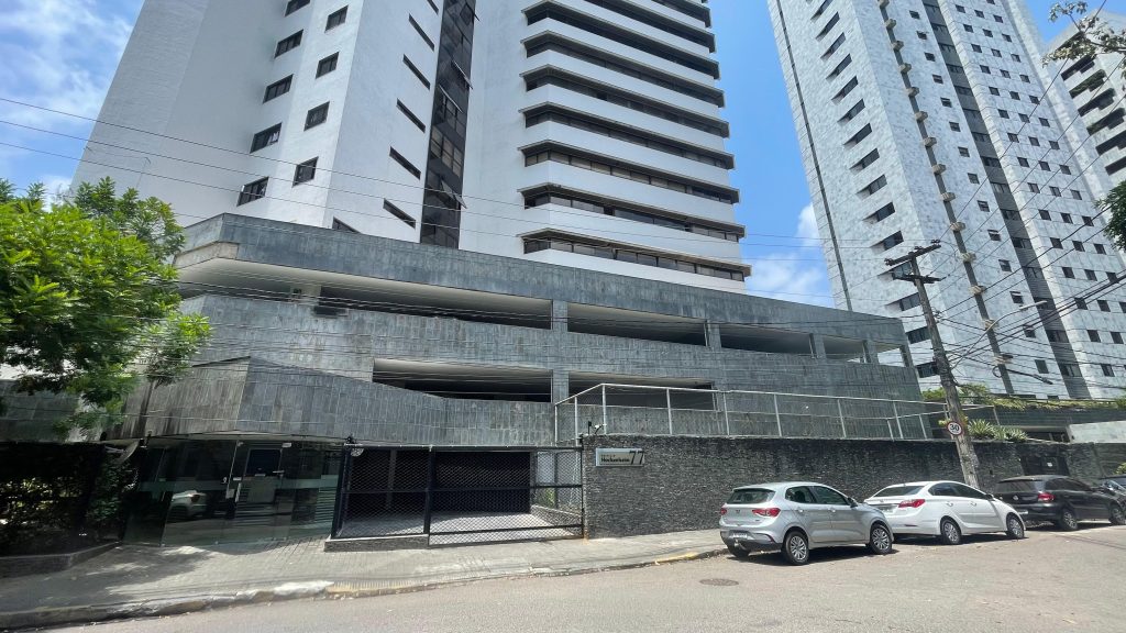 A foto mostra a fachada de um prédio residencial alto, com garagem em vários níveis e carros estacionados na rua em frente.