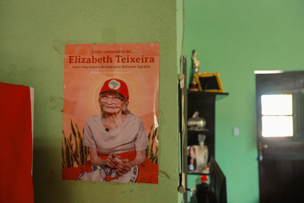 A foto mostra um cartaz fixado em uma parede verde, em homenagem ao centenário de Elizabeth Teixeira, figura histórica da luta pela Reforma Agrária no Brasil. No cartaz, ela aparece aos 100 anos, sentada, com expressão serena, usando boné vermelho do MST, óculos, blusa clara e uma saia com símbolos do movimento. O texto diz: “2025: centenário de Elizabeth Teixeira – Uma vida inteira de luta pela Reforma Agrária. Homenagem de toda militância do MST”.