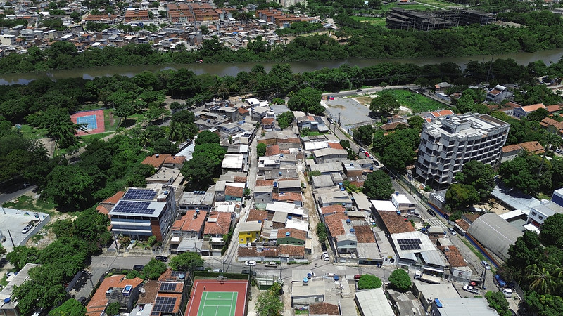 A foto mostra uma vista aérea da comunidade de Santana, no Recife. As casas estão dispostas de forma densa e irregular, com telhados de cerâmica em tons terrosos. À esquerda, aparece uma área verde com árvores frondosas que contrasta com a urbanização. Também se destaca uma quadra poliesportiva colorida, cercada por grades, usada pela comunidade. No canto inferior direito, há uma rua mais larga com edificações maiores e outras árvores. Ao fundo, vê-se o entorno da comunidade, com mais vegetação e construções espalhadas. A cena transmite a ideia de forte adensamento urbano combinado com espaços de convivência e lazer.