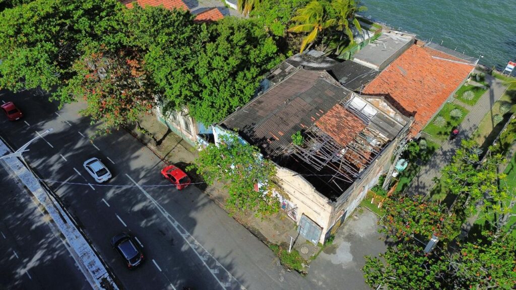 A imagem aérea mostra um prédio antigo e parcialmente degradado, visto de cima. O telhado está danificado, com uma parte destruída que revela estrutura metálica exposta e ausência de cobertura. Ao redor, há vegetação abundante com árvores cujas copas avançam sobre parte da edificação. À esquerda, observa-se uma rua com alguns carros estacionados e faixa de pedestre. À direita, nota-se a proximidade de um corpo d’água, evidenciado por um trecho de calçada verde com pequeno jardim e piso regular.