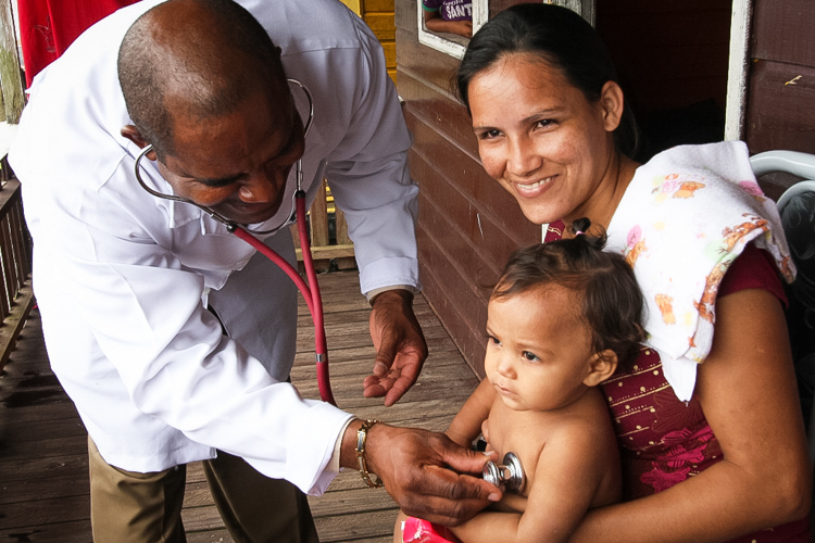 Na foto, um médico negro está examinando uma criança pequena com um estetoscópio. Ele está inclinado para frente, sorridente e concentrado, ouvindo o coração da criança. A criança está sentada no colo de uma mulher, provavelmente a mãe, que sorri para a câmera. A criança aparece sem camisa para permitir o exame. A mãe veste uma blusa vermelha estampada e tem um pano branco apoiado no ombro. A cena acontece em um ambiente simples, com paredes de madeira, dando a impressão de uma casa ou posto de saúde comunitário.