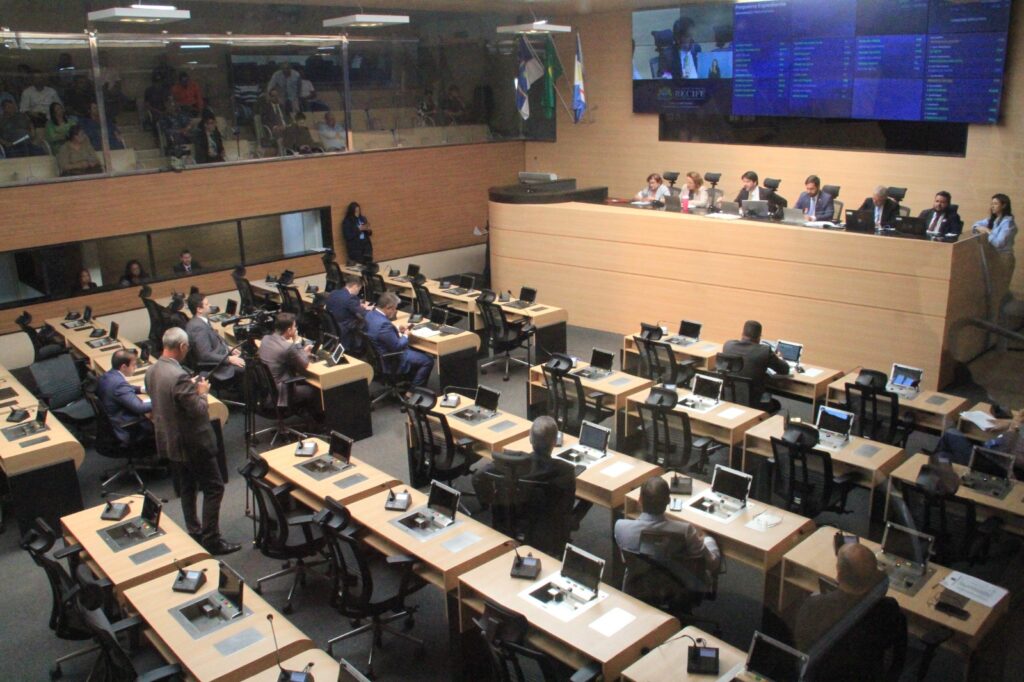 A foto mostra o plenário da câmara de vereadores em funcionamento. O espaço é amplo, moderno e bem iluminado, com várias mesas individuais organizadas em fileiras, cada uma equipada com microfones e telas. Alguns vereadores estão sentados em suas cadeiras, enquanto outros circulam pelo ambiente. Na parte da frente, em um nível mais elevado, há uma mesa maior onde ficam os membros da mesa diretora, homens e mulheres, acompanhando e conduzindo a sessão. Acima deles, há telas eletrônicas grandes que exibem informações da votação ou da pauta do dia. Ao fundo, no mezanino, estão alguns cidadãos assistindo à sessão.