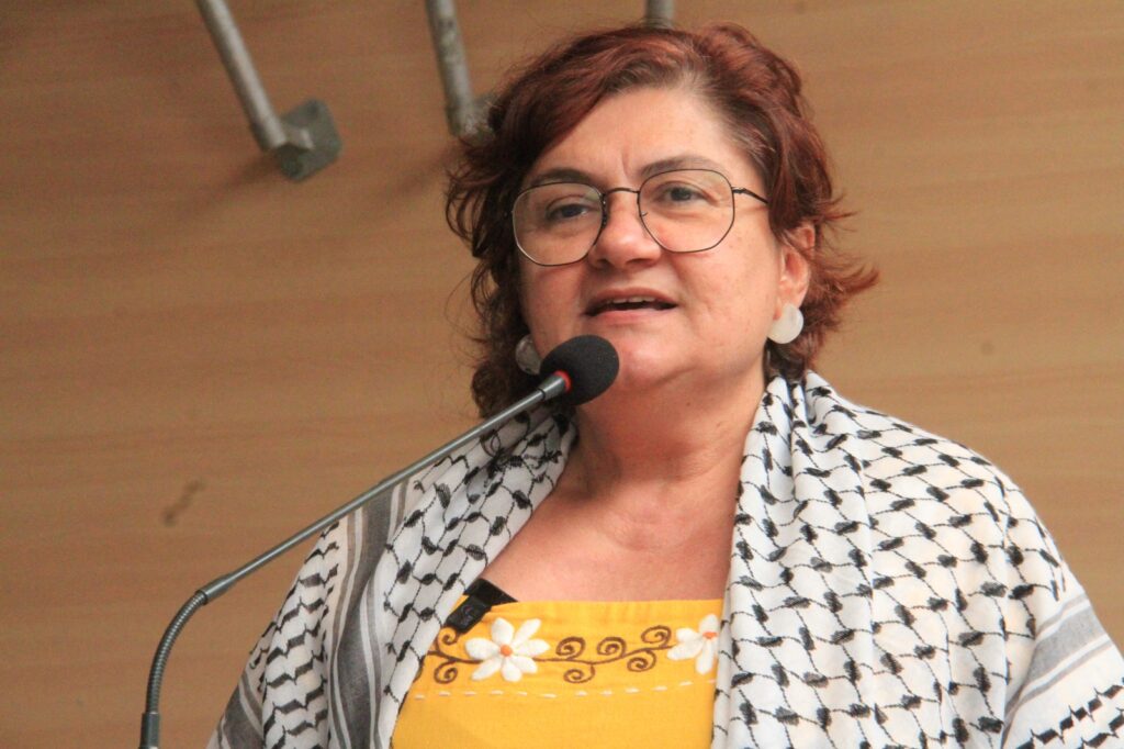 A foto mostra a vereadora Cida Pedrosa falando ao microfone em um ambiente fechado, provavelmente durante uma sessão ou evento oficial. Ela está em pé, com expressão séria, como se estivesse discursando. Usa óculos de aro fino, brincos arredondados claros, um vestido amarelo com bordados florais brancos sob um lenço palestino estampado em preto e branco sobre os ombros. O fundo é liso, de madeira clara, destacando ainda mais a figura dela em primeiro plano.