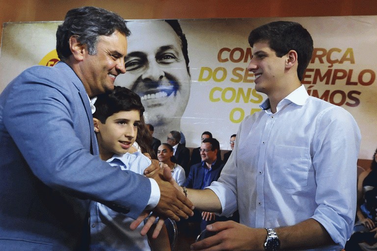 Na foto, João Campos, de camisa branca, sorri enquanto aperta a mão de Aécio Neves, que usa paletó azul claro. Um menino observa a cena entre os dois. Ao fundo, há um grande banner com o rosto de Eduardo Campos e parte de uma frase em destaque, além de pessoas sentadas.