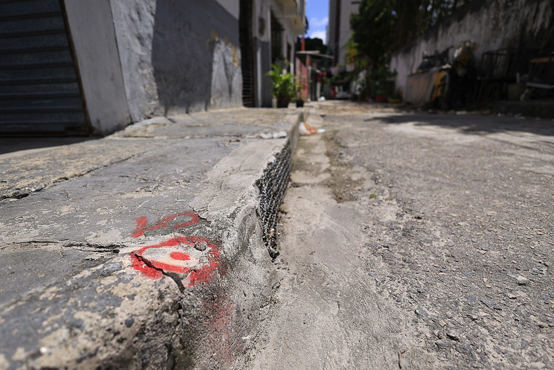A foto mostra uma rua estreita e pavimentada da comunidade de Santana, vista em perspectiva baixa, bem próxima ao chão. Em primeiro plano, aparece uma calçada desgastada e quebrada, com rachaduras no cimento. Nela há uma marca de tinta vermelha em formato circular, feita de forma improvisada, como uma espécie de sinalização. O meio-fio também está danificado, com parte exposta de ferro ou arame. Mais ao fundo, a rua segue entre casas e muros, com algumas plantas em vasos e objetos espalhados. A cena transmite a ideia de descuido na infraestrutura e chama atenção para as marcas vermelhas, que sugerem algum tipo de intervenção planejada.