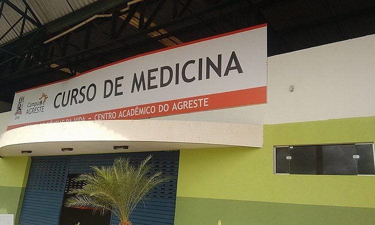 A foto mostra a fachada de um prédio com uma grande placa branca e faixa vermelha, onde se lê em letras maiúsculas: “CURSO DE MEDICINA”. Acima, aparece o logotipo da Universidade Federal de Pernambuco e a identificação “Campus do Agreste”. Abaixo da faixa, há uma parede pintada em verde e amarelo-claro, com portas de enrolar azuis fechadas. Na frente do prédio, vê-se uma pequena palmeira decorativa.
