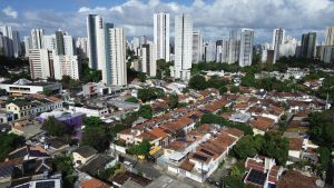A foto aérea mostra uma parte da zona norte do Recife, em Pernambuco. Em primeiro plano, aparecem várias casas térreas e sobrados com telhados de cerâmica vermelha, muitas delas equipadas com placas solares. As ruas são estreitas e arborizadas, formando um bairro de aspecto residencial tradicional. Ao fundo, a paisagem muda radicalmente: ergue-se uma fileira densa de prédios altos e modernos, de cores claras, característicos da verticalização da cidade. O contraste entre as construções baixas, antigas e coloridas, e os arranha-céus contemporâneos é marcante. No céu, nuvens brancas se espalham sob a luz do sol, reforçando a sensação de um dia claro.