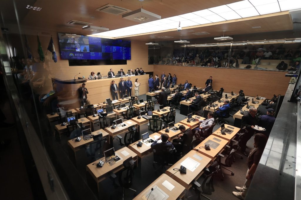 A foto mostra o plenário da Câmara Municipal do Recife durante uma sessão. O espaço é amplo, com mesas de madeira clara organizadas em fileiras, cada uma equipada com microfones, computadores e cadeiras pretas. Vários vereadores estão sentados, alguns conversam em pé, e outros circulam pelo ambiente. Na parte frontal, sobre uma bancada elevada, estão os membros da mesa diretora. Atrás deles, uma grande tela digital exibe informações da sessão e imagens de vereadores. À esquerda, bandeiras ficam expostas. Na parte superior, atrás de um vidro, há uma galeria com público acompanhando os trabalhos. A iluminação é clara e uniforme, destacando o ambiente moderno e organizado.