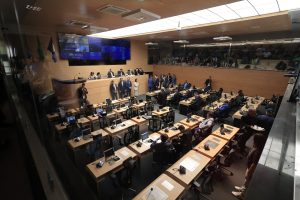 A foto mostra o plenário da Câmara Municipal do Recife durante uma sessão. O espaço é amplo, com mesas de madeira clara organizadas em fileiras, cada uma equipada com microfones, computadores e cadeiras pretas. Vários vereadores estão sentados, alguns conversam em pé, e outros circulam pelo ambiente. Na parte frontal, sobre uma bancada elevada, estão os membros da mesa diretora. Atrás deles, uma grande tela digital exibe informações da sessão e imagens de vereadores. À esquerda, bandeiras ficam expostas. Na parte superior, atrás de um vidro, há uma galeria com público acompanhando os trabalhos. A iluminação é clara e uniforme, destacando o ambiente moderno e organizado.