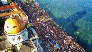 A foto mostra uma grande manifestação na Rua da Aurora, no Recife, vista de cima. À esquerda, aparece a cúpula dourada de um prédio histórico, que se destaca no primeiro plano. A rua, bem larga e paralela ao Rio Capibaribe, está completamente tomada por uma multidão compacta de milhares de pessoas. A massa de manifestantes ocupa toda a via até onde a vista alcança, criando um mar de gente. Muitas pessoas carregam bandeiras, faixas e balões, que aparecem distribuídos no meio da multidão, adicionando pontos de cor. À direita da imagem, o rio reflete o céu, com sombras longas projetadas sobre a água. Na rua, mais próxima ao rio, há carros de som e trios elétricos, de onde provavelmente partem discursos e músicas para o protesto.