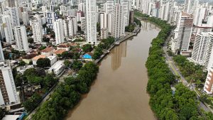 Vista aérea do Recife com muitos prédios altos dos dois lados do Rio Capibaribe, na altura dos bairros das Graças e Torre. As margens do rio são cobertas por árvores verdes e há uma avenida movimentada à direita da imagem