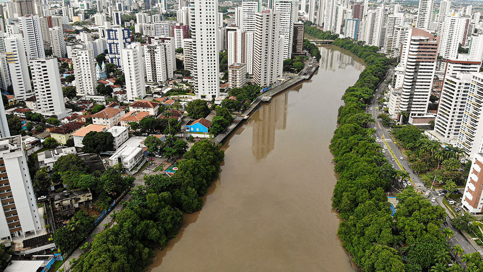 Vista aérea do Recife com muitos prédios altos dos dois lados do Rio Capibaribe, na altura dos bairros das Graças e Torre. As margens do rio são cobertas por árvores verdes e há uma avenida movimentada à direita da imagem