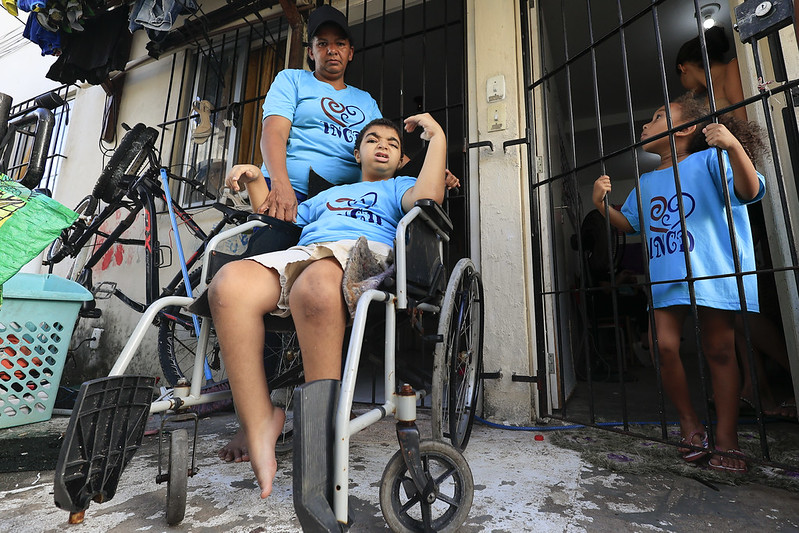 A foto mostra uma mulher e duas crianças em frente a uma casa simples, com grades de ferro nas portas. A mulher, de expressão firme, veste camiseta azul-clara com um logotipo vermelho em forma de coração estilizado com as letras INCD abaixo. Ela está atrás de um menino em cadeira de rodas, também com a mesma camiseta. Ele está descalço e com uma das pernas apoiada sobre o descanso da cadeira. À direita, uma menina observa a cena de dentro da casa, segurando a grade, igualmente vestida com a camiseta azul. Ao lado da casa há uma bicicleta encostada e uma cesta plástica. O chão é de cimento gasto, e o ambiente sugere um cotidiano modesto, marcado por afeto, cuidado e resistência familiar.