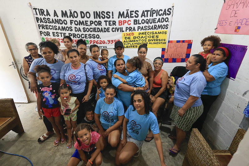 A foto mostra cerca de 20 mulheres reunidas em um salão iluminado, posando em grupo. A maioria veste camisetas azuis e roupas casuais, e uma delas segura uma criança. Ao fundo, há um grande banner branco com frases de protesto em letras coloridas, pedindo respeito e apoio a “mães atípicas” e denunciando bloqueios de benefícios do INSS. As participantes demonstram união e firmeza, com expressões sérias ou discretamente sorridentes. Algumas se apoiam em cadeiras ou nos ombros umas das outras. O ambiente tem piso claro, paredes brancas e luz intensa, sugerindo um encontro ou mobilização social por direitos e assistência.