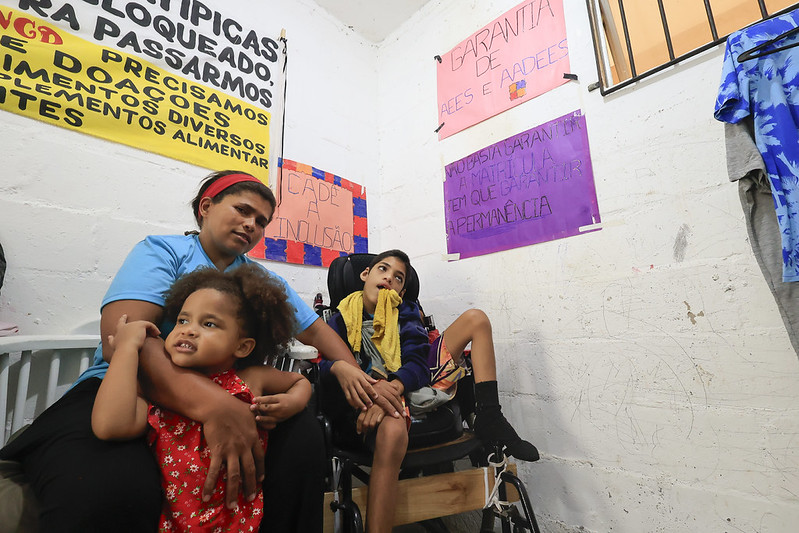 A foto mostra uma mulher sentada em um quarto simples, de paredes brancas, segurando uma menina pequena no colo. Ao lado delas está um menino em cadeira de rodas, com expressão tranquila e uma toalha amarela sobre o ombro. A mulher veste camiseta azul e calça escura; a menina, um vestido vermelho estampado. O ambiente é iluminado e tem cartazes coloridos nas paredes com mensagens escritas à mão, pedindo inclusão e garantia de direitos para pessoas com deficiência. Há também um varal com roupas e sinais de uso cotidiano nas paredes. A imagem transmite afeto, cuidado e a luta por dignidade e apoio social.