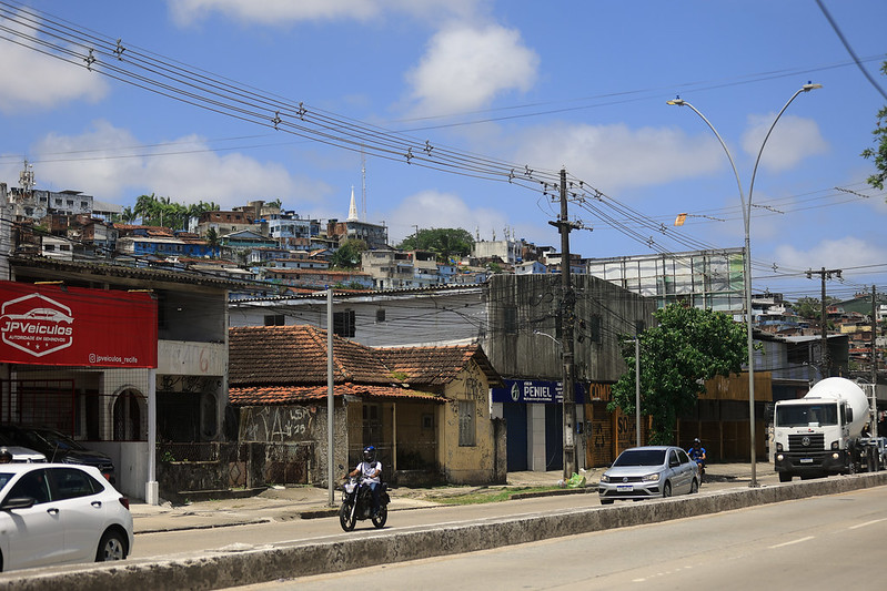 A foto mostra uma avenida movimentada em um bairro urbano. Na pista, passam um caminhão branco, um carro prata e uma motocicleta conduzida por um homem de capacete. À direita e à esquerda da via há calçadas, postes e fios de energia. As construções ao longo da rua são simples, com fachadas desgastadas e coberturas de telha, abrigando pequenos comércios, como uma loja chamada “JP Veículos” e outra com o letreiro “Peniel”. Ao fundo, em uma encosta, aparecem muitas casas amontoadas, formando uma comunidade densa, e ao centro do horizonte se destaca a ponta branca de uma torre de igreja. O céu está azul com algumas nuvens, e a luz do sol é forte, indicando um dia claro e quente.