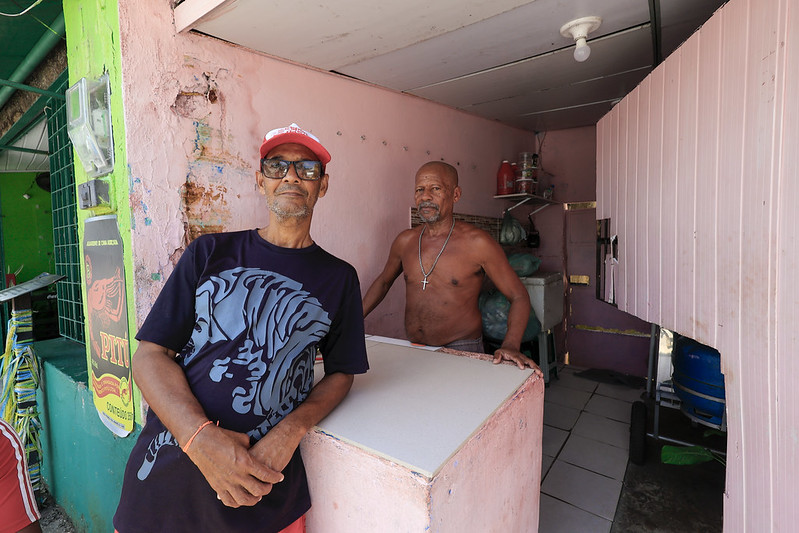 A foto mostra dois homens em um pequeno comércio de paredes pintadas de rosa. O homem à esquerda está de pé, encostado em um balcão, usando boné vermelho, óculos escuros e camiseta preta com estampa de tigre. Ele olha para a câmera com expressão calma. O homem à direita está sem camisa, usa um cordão com crucifixo e está atrás do balcão, dentro do espaço do comércio, onde há prateleiras com garrafas e sacos plásticos empilhados. A iluminação natural vem da rua, destacando os tons claros das paredes e do piso branco.