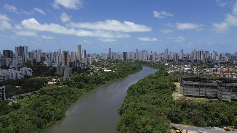 Foto aérea do Recife mostra o Capibaribe, um rio sinuoso cortando a paisagem urbana. As margens do rio são cobertas por densa vegetação verde, contrastando com a cidade ao redor. À esquerda e ao fundo, ergue-se uma grande concentração de prédios altos e modernos; à direita, aparecem áreas com construções mais baixas e um prédio em obras. O céu azul, com nuvens brancas espalhadas, completa a cena, transmitindo uma sensação de dia ensolarado e quente típico do clima tropical.