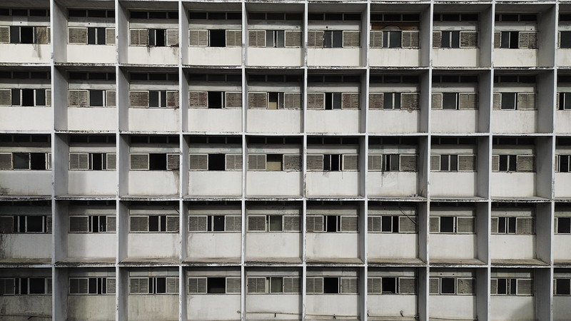 O ChatGPT disse: Foto mostra a fachada de um edifício antigo e desgastado, composta por várias janelas dispostas em fileiras e colunas regulares, formando um padrão geométrico repetitivo. As paredes são brancas, mas apresentam manchas de sujeira, descascados e sinais de deterioração. Algumas janelas estão abertas, outras fechadas, e várias têm venezianas de madeira envelhecidas ou danificadas. A imagem transmite uma sensação de abandono e desgaste urbano.