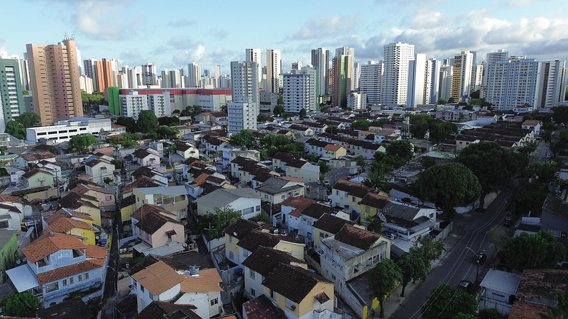 Foto aérea da Vila dos Comerciários, na zona norte do Recife, mostra um conjunto de casas baixas e próximas umas das outras, com telhados de cerâmica em tons de marrom e vermelho. As ruas são estreitas e arborizadas, e há árvores de copa larga espalhadas entre as residências. Ao fundo, ergue-se uma extensa paisagem de prédios altos e modernos, formando um contraste marcante entre o bairro tradicional e o crescimento vertical da cidade. O céu está parcialmente nublado, com luz suave iluminando a cena urbana.