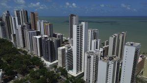 O ChatGPT disse: Foto aérea do bairro de Boa Viagem, no Recife, mostrando uma fileira de prédios altos e modernos à beira-mar. As construções, em sua maioria de tons claros como branco, bege e cinza, formam uma paisagem vertical que se estende paralela à orla. À esquerda, há uma faixa verde com árvores que acompanha uma avenida larga. Ao fundo, o mar aparece em tons de verde e azul, com o horizonte se unindo ao céu parcialmente nublado. A imagem transmite a sensação de urbanização intensa junto à costa